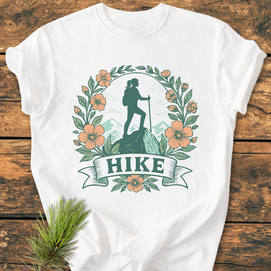 HIKE Flower - Vintage Wash Tee features a stylized hiker design with mountain backdrop, framed by leaves and flowers, on a white T-shirt laid on wood, accompanied by a pine sprig.