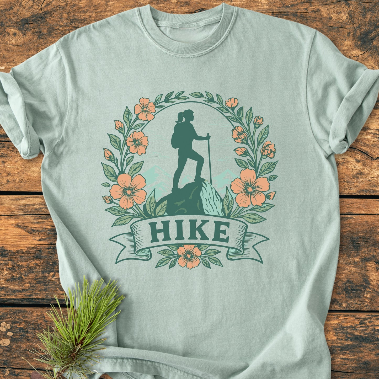 HIKE Flower - Vintage Wash Tee features a nature-themed design with a backpacking hiker and mountain silhouette, encircled by orange flowers and green leaves, on a light-mint, soft-washed garment-dyed cotton fabric.