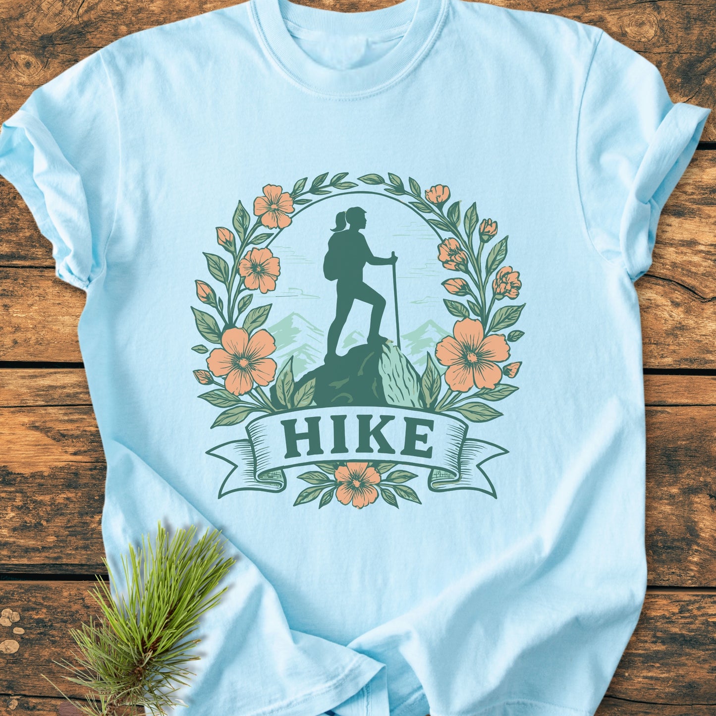 The HIKE Flower - Vintage Wash Tee features a hiker silhouette with a mountain backdrop, encircled by leaves and flowers, printed on a light-blue, 100% ring-spun cotton T-shirt.