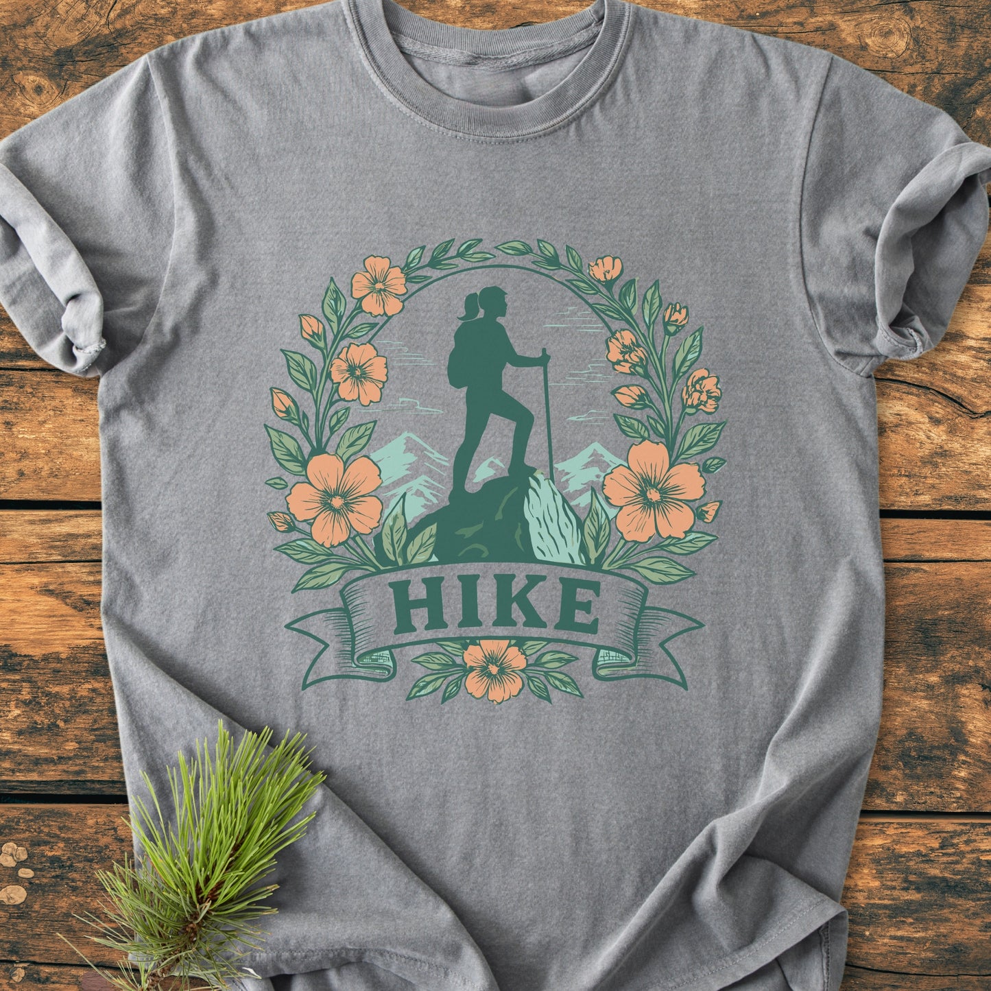 Gray HIKE Flower - Vintage Wash Tee featuring a hiking silhouette framed by leaves and flowers. Laid flat, highlighting the soft, relaxed fit and durable stitching. Pine sprig accentuates the natural theme.