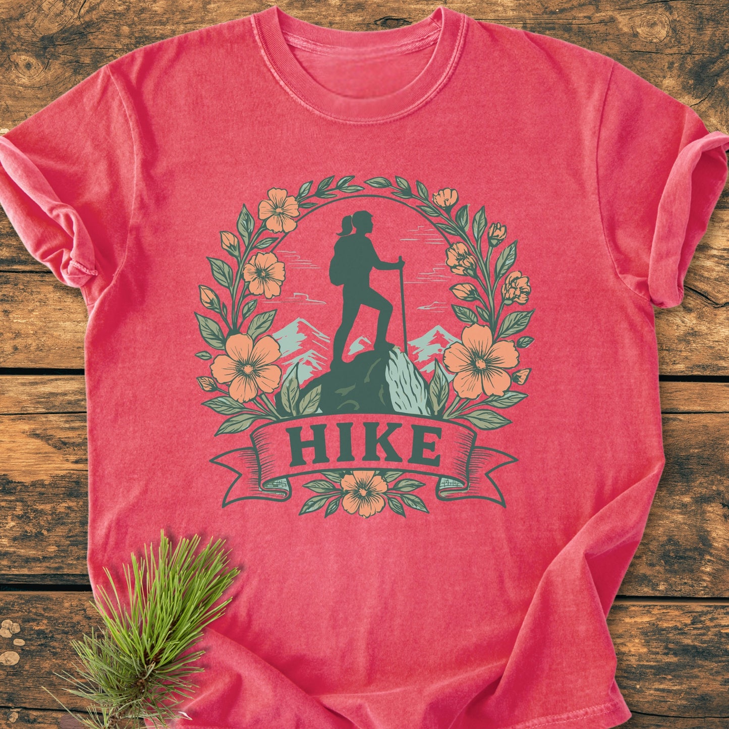HIKE Flower - Vintage Wash Tee: Heather-rose T-shirt featuring a hiking silhouette amid mountain peaks, encircled by leaves and flowers, with HIKE on a ribbon. Laid on wood with pine sprig.