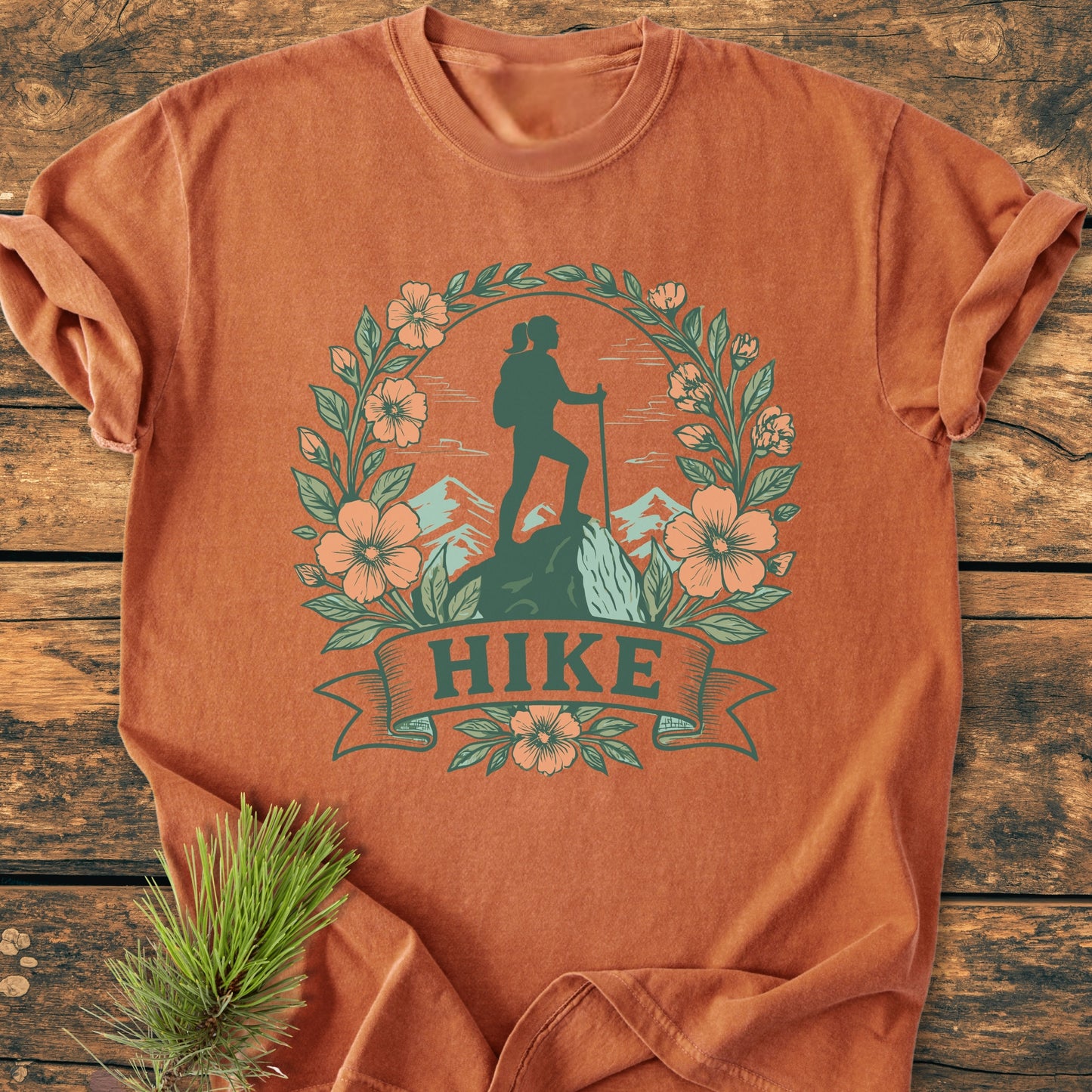 HIKE Flower - Vintage Wash Tee features a hiker graphic with mountains and flowers, laid on a wooden surface. A pine sprig accents this soft, durable, 100% cotton, relaxed fit T-shirt.