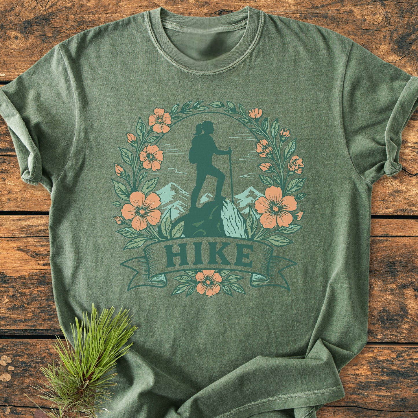 Heather-green HIKE Flower - Vintage Wash Tee with a hiker silhouette and leafy wreath printed on the front, laid on rustic wood, featuring a pine sprig accent.