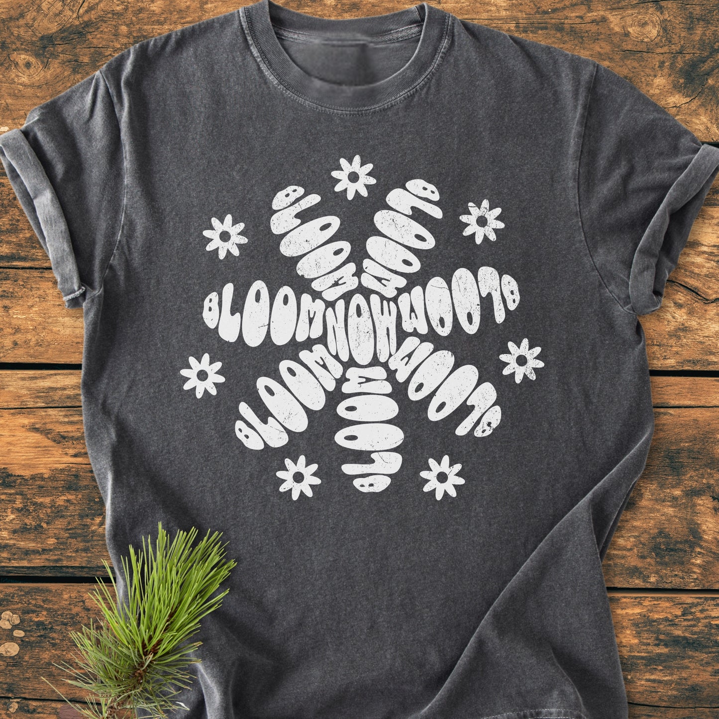 Bloom Now - Vintage Wash Tee features a charcoal-gray T-shirt with a white floral graphic, laid on wood. The soft, relaxed fit tee is made of pre-shrunk, ring-spun cotton, emphasizing durability.