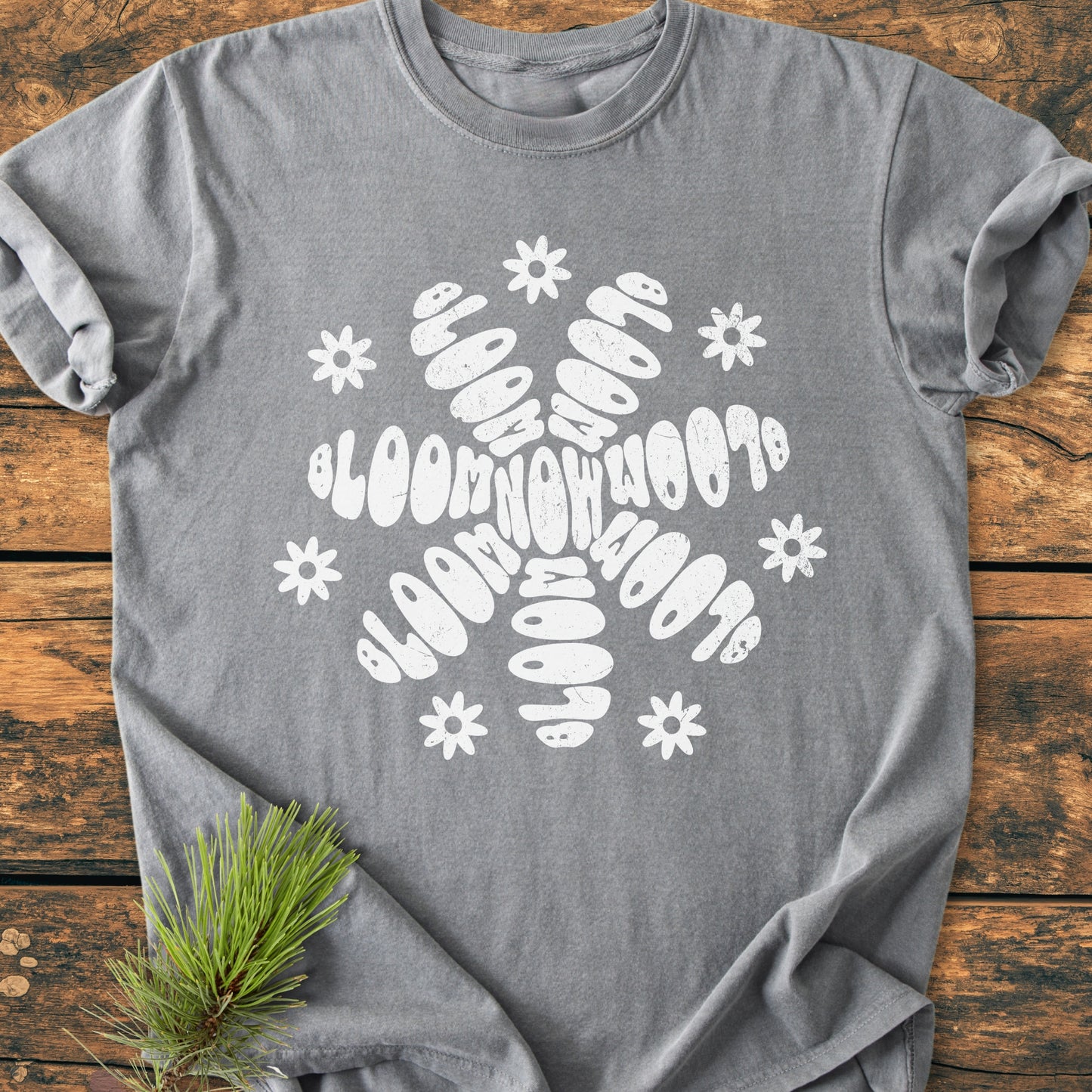 Bloom Now - Vintage Wash Tee: A heather-gray T-shirt with a white floral BLOOM motif, featuring daisy accents, laid on wood. Pine sprig decorates the scene, highlighting the tee's relaxed, cozy fit.
