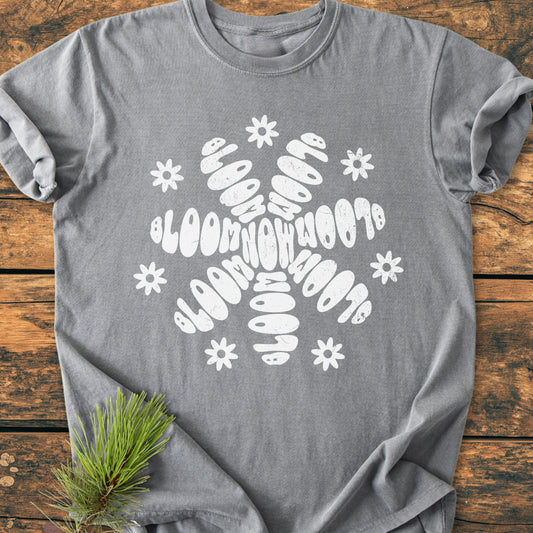 Bloom Now - Vintage Wash Tee: A heather-gray T-shirt with a white floral BLOOM motif, featuring daisy accents, laid on wood. Pine sprig decorates the scene, highlighting the tee's relaxed, cozy fit.