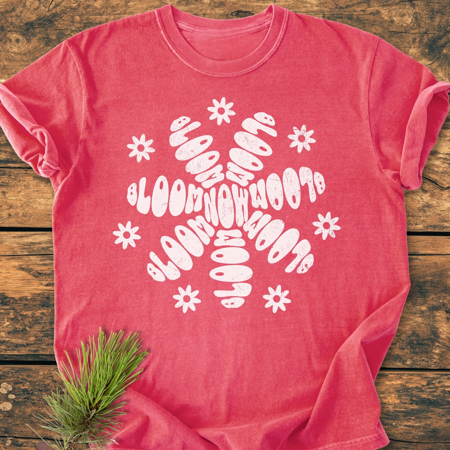Bloom Now - Vintage Wash Tee in faded-coral features a white stylized flower/snowflake graphic with bloom now phrases and daisy motifs, laid on a rustic wooden surface with a pine needle sprig.