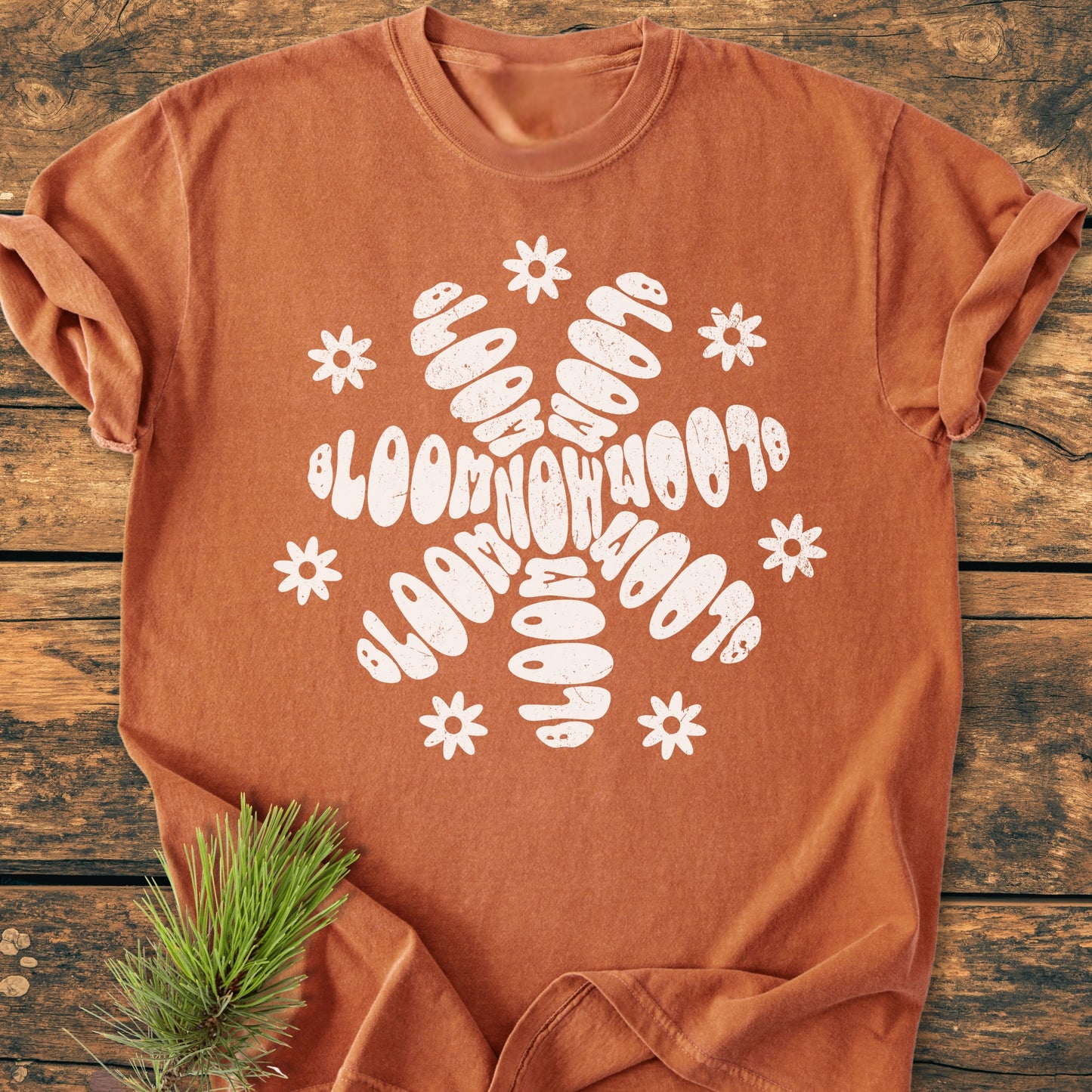 Bloom Now - Vintage Wash Tee features a retro-psychedelic snowflake motif with bubble-letter arms and daisy flowers. The rust-orange T-shirt lies flat, showcasing its relaxed fit and soft cotton fabric.