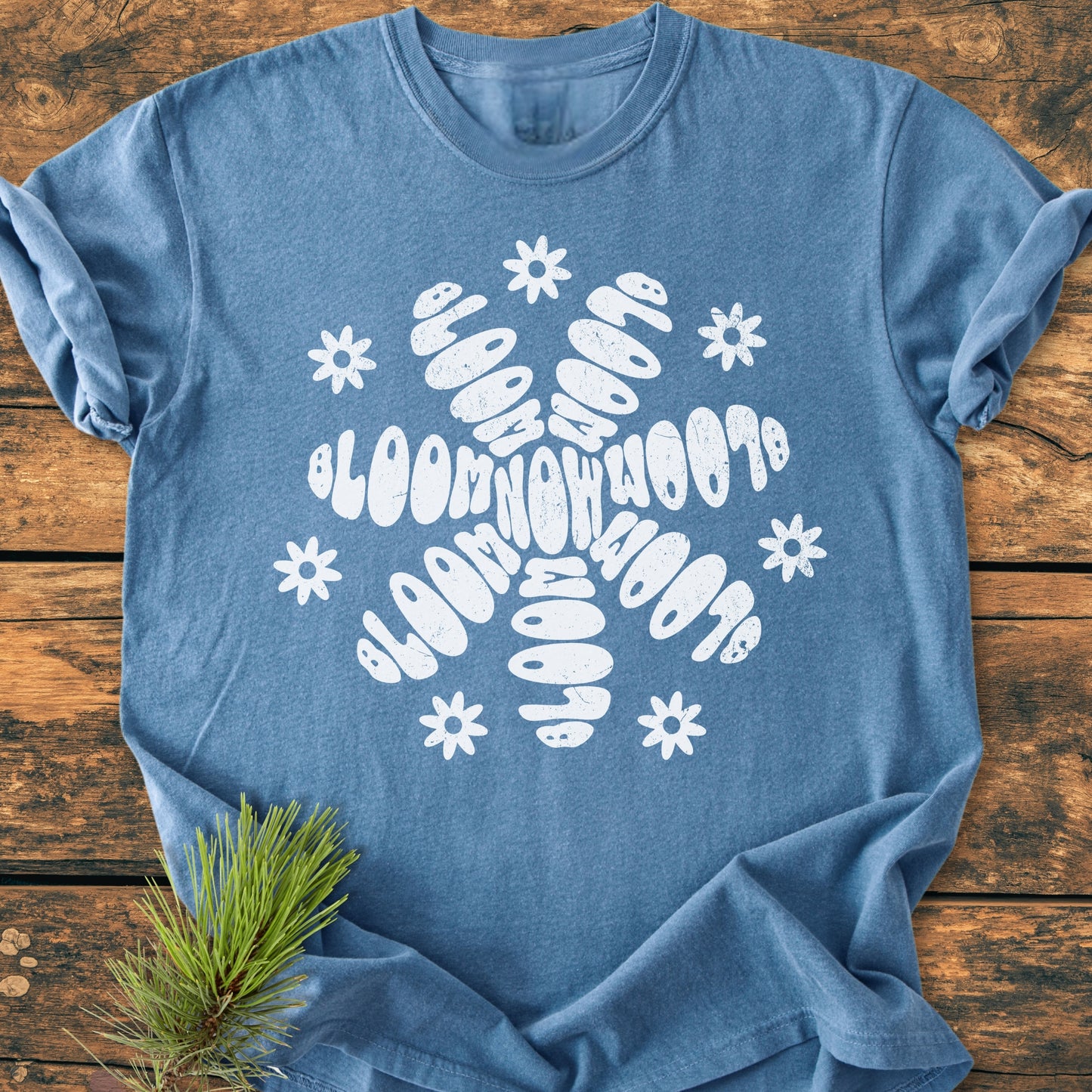 Bloom Now - Vintage Wash Tee: A heather-blue T-shirt with a white snowflake graphic featuring BLOOM NOW 2018 in bubble letters, accented by small daisies, laid on a wooden surface.