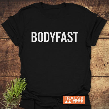 Black BODYFAST T-Shirt with bold white lettering, styled on wood with pine sprig. Features TRAILS & TEES logo, symbolizing an energized lifestyle inspired by the BodyFast app.