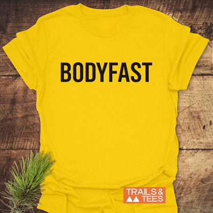 Bright yellow BODYFAST T-Shirt with bold black lettering, laid flat on wood. Features TRAILS & TEES logo on the side and a pine sprig accent in the corner.