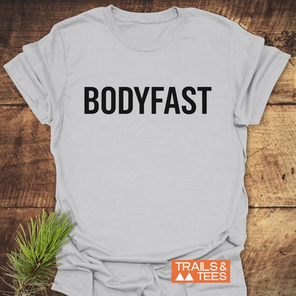 Light-colored BODYFAST T-Shirt with bold black lettering on the chest, styled on a wooden surface with a pine sprig, symbolizing energy and balance from the BodyFast lifestyle.