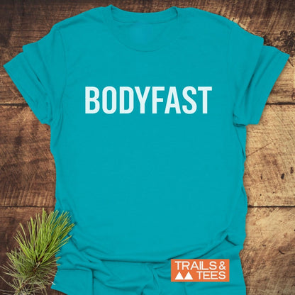 BODYFAST T-Shirt displayed on a wooden surface, features bold white lettering. Accompanied by an orange TRAILS & TEES logo with mountain icons, and a pine sprig accent.