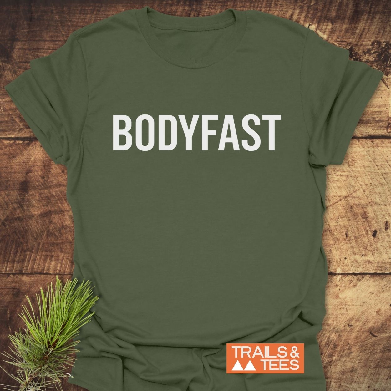 Olive-green T-shirt with BODYFAST in bold white letters, representing a lifestyle of energy and balance. Includes TRAILS & TEES logo and pine sprig accent on rustic wood background.
