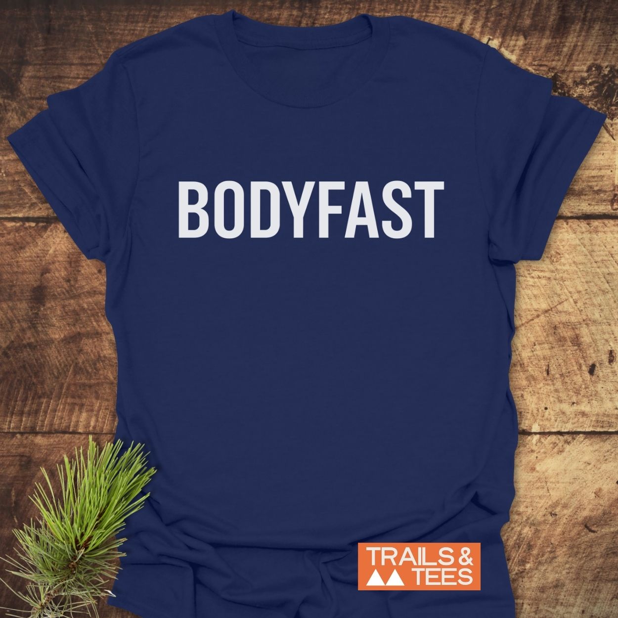 Navy-blue BODYFAST T-Shirt displayed on wood, featuring bold white lettering and a small orange “TRAILS & TEES” logo, with pine needles adding a natural touch beside the shirt.