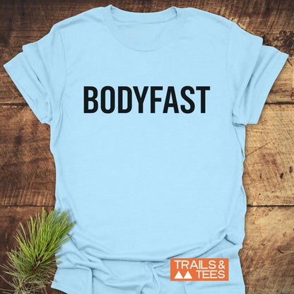 Light-blue BODYFAST T-Shirt with bold black lettering, laid flat on wood. Includes “TRAILS & TEES” orange label. Pine sprig accentuates the lifestyle theme of energy and balance.