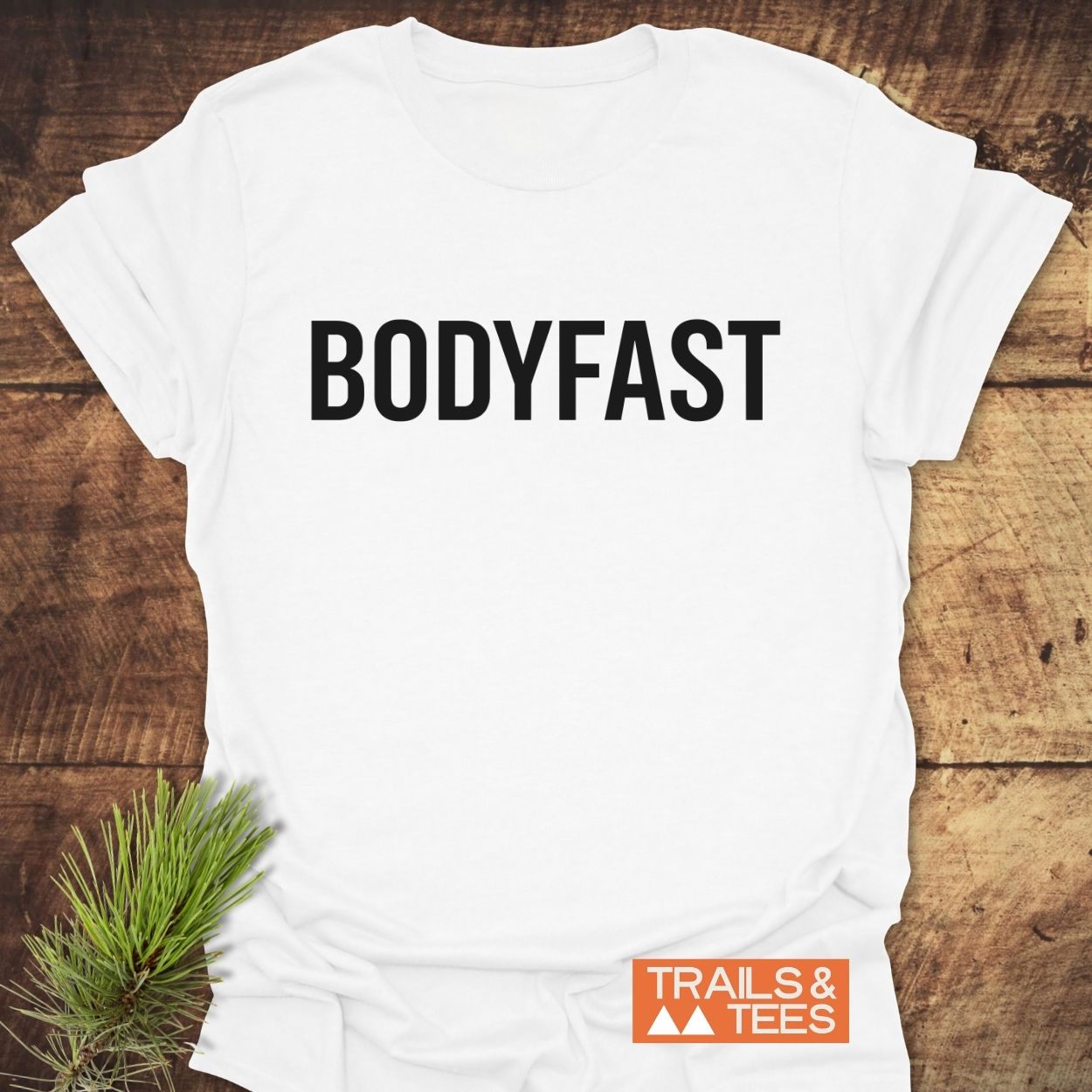 BODYFAST T-Shirt featuring bold black lettering across the chest, with a “TRAILS & TEES” orange label in the corner, symbolizing an energized lifestyle, laid on a wooden surface.