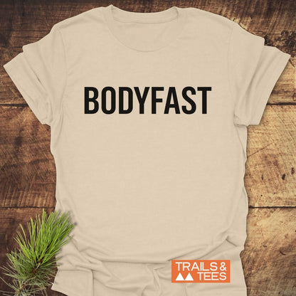 Light-beige BODYFAST T-Shirt on rustic wooden surface, featuring bold black lettering. Includes “TRAILS & TEES” logo and pine sprig, symbolizing an active, balanced lifestyle.