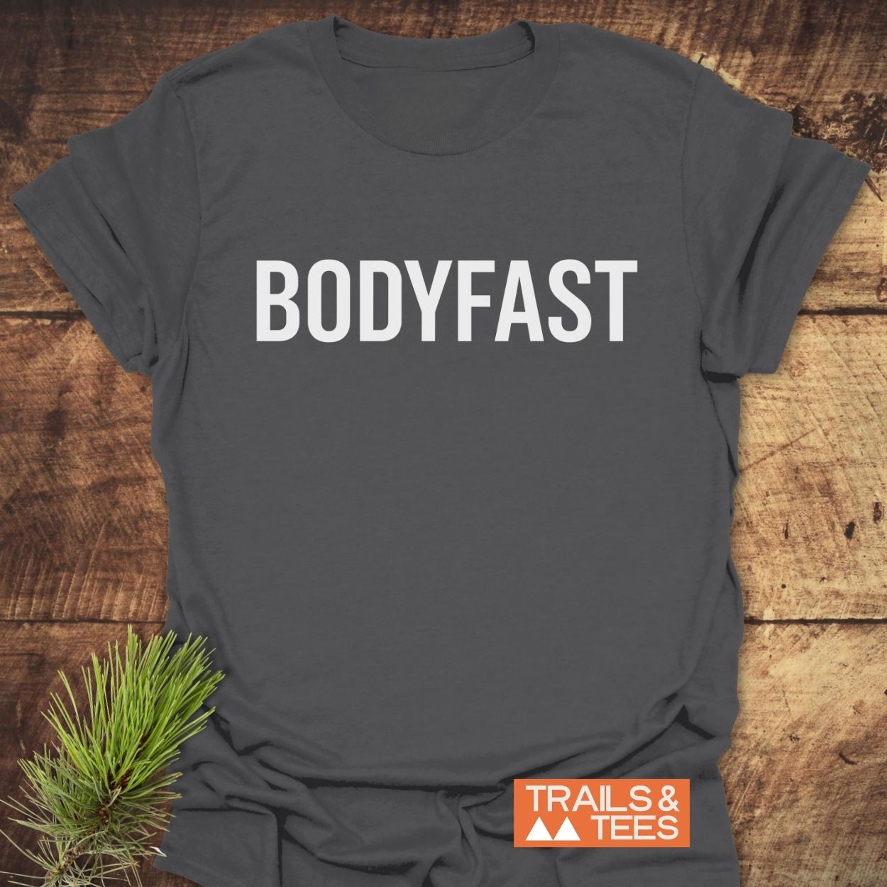 Dark-gray BODYFAST T-Shirt on rustic wood surface, featuring bold white lettering across the chest. Includes TRAILS & TEES orange logo with mountain design in corner, accompanied by a pine sprig.