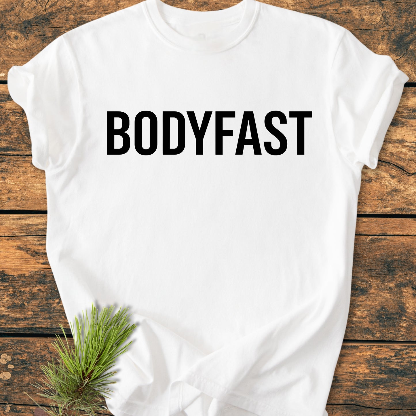 BODYFAST - Vintage Wash Tee featuring BODYFAST in bold black letters on a white T-shirt, laid on rustic wood with a pine sprig, symbolizing an energized, balanced lifestyle.