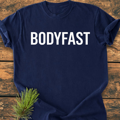 Navy blue BODYFAST - Vintage Wash Tee with bold white lettering, laid flat on wooden surface. A small pine sprig adds a natural touch, symbolizing the balanced, energetic lifestyle BODYFAST promotes.