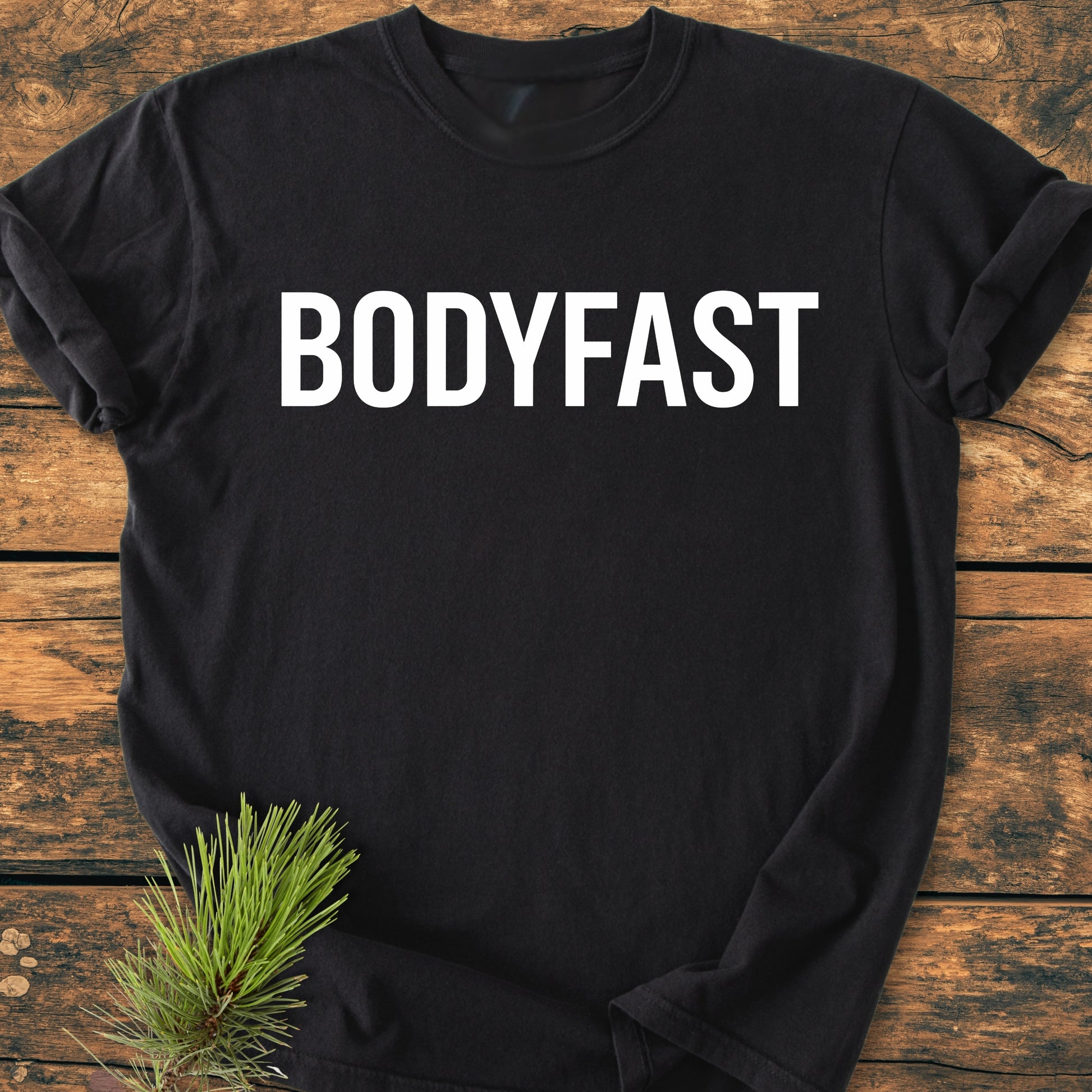 BODYFAST - Vintage Wash Tee on a rustic wooden surface, featuring bold white BODYFAST lettering. Pine needles add a natural touch, emphasizing the lifestyle of energy and balance.
