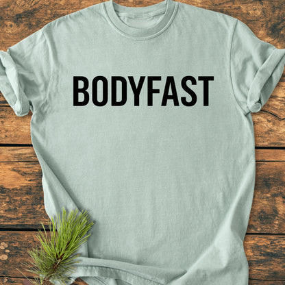 BODYFAST - Vintage Wash Tee featuring bold black lettering on the chest, laid flat on a rustic wood surface with a pine needle sprig in the corner, symbolizing an energized lifestyle.
