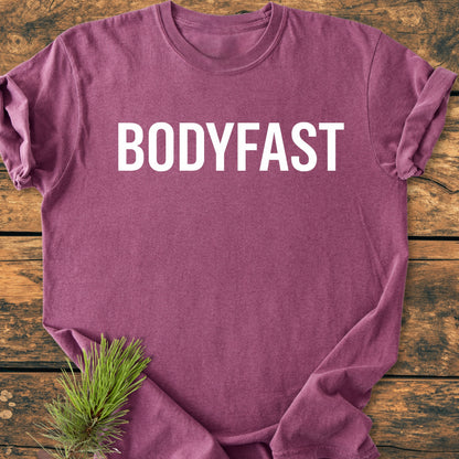 Heathered plum BODYFAST - Vintage Wash Tee on rustic wood surface, bold white BODYFAST text on chest, accented by pine sprig, embodying the app’s lifestyle of energy and balance.