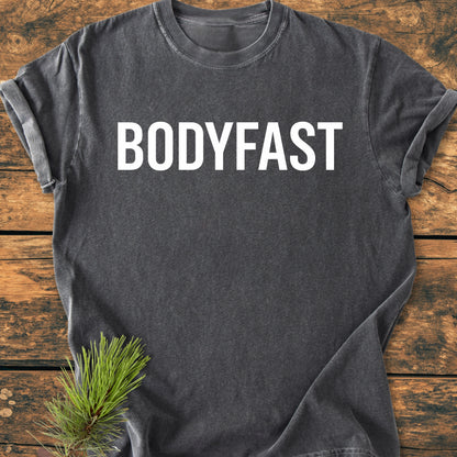 Charcoal-gray BODYFAST - Vintage Wash Tee on rustic wood, featuring bold white logo across chest with a pine sprig accent, symbolizing a balanced, energetic lifestyle promoted by the BodyFast app.