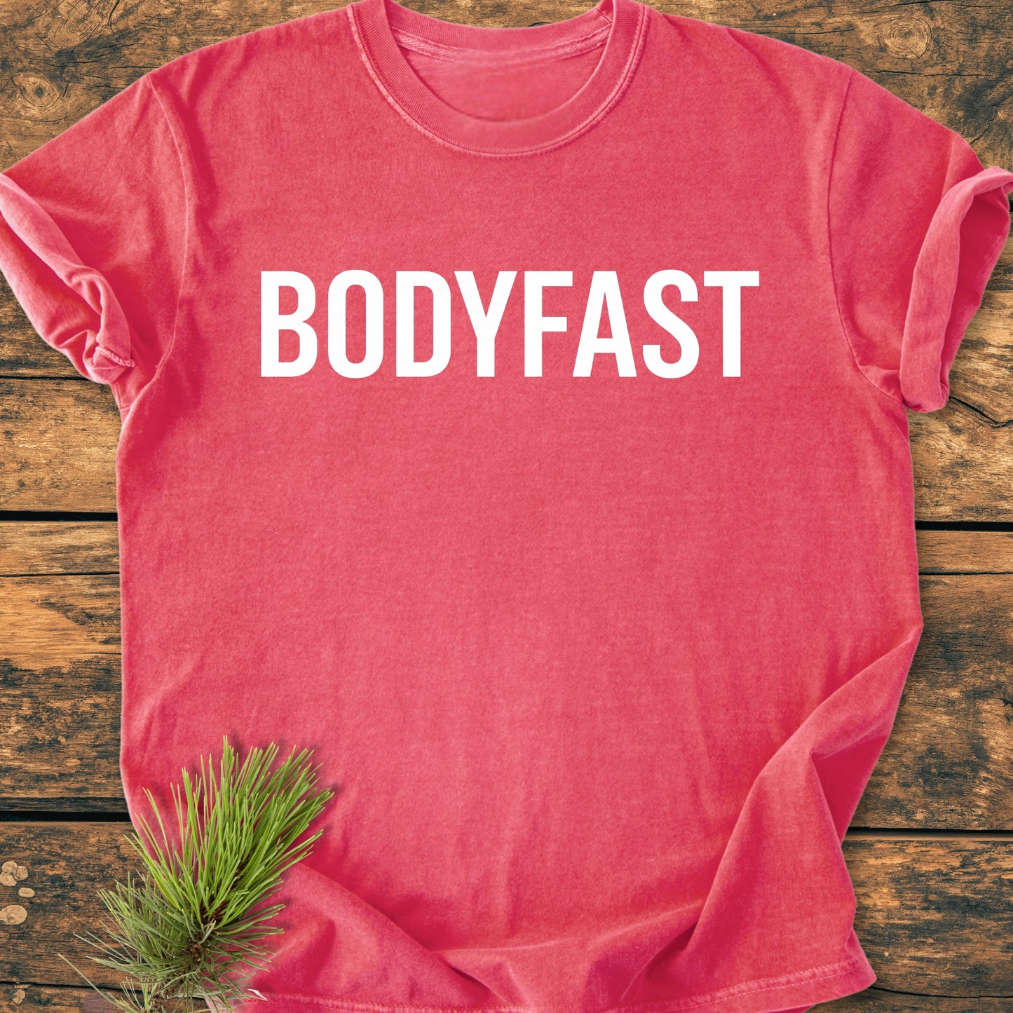 The BODYFAST - Vintage Wash Tee features bold BODYFAST lettering across the chest, laid flat on a wooden surface, with a pine sprig accentuating its lifestyle-inspired design.