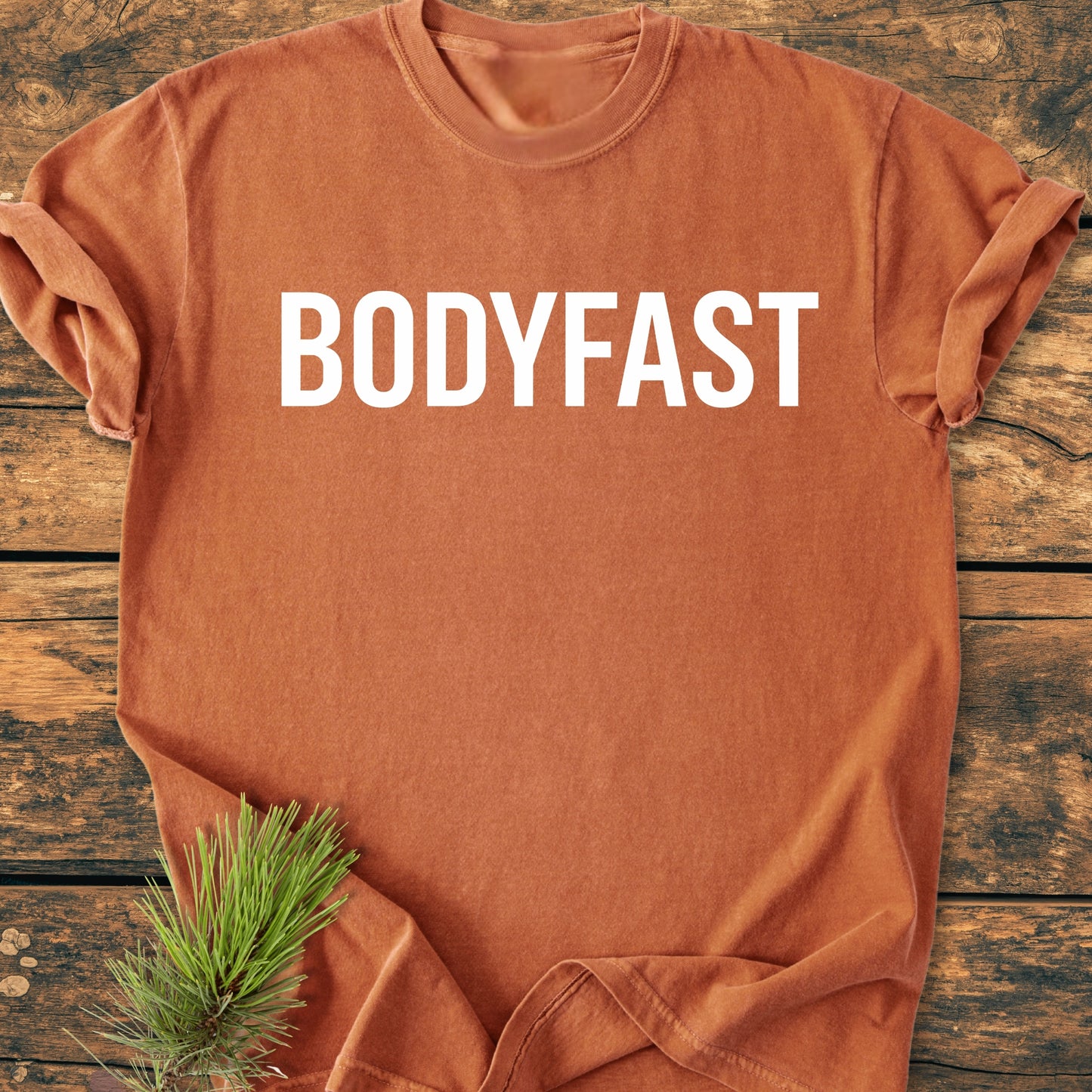 Rust-orange BODYFAST - Vintage Wash Tee with bold white lettering on chest, displayed flat on wood surface; pine sprig adds natural accent, embodying an active, balanced lifestyle.