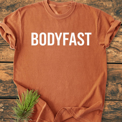 Rust-orange BODYFAST - Vintage Wash Tee with bold white lettering on chest, displayed flat on wood surface; pine sprig adds natural accent, embodying an active, balanced lifestyle.
