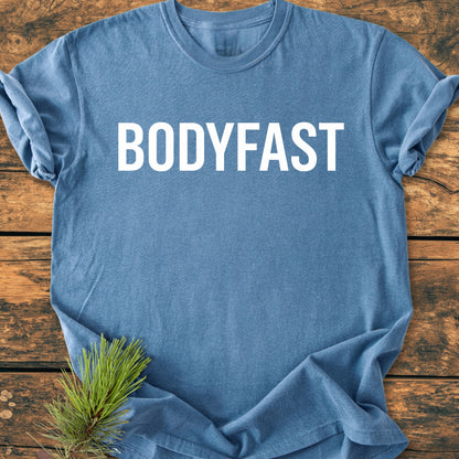 BODYFAST - Vintage Wash Tee, featuring bold white lettering across the chest, laid on wood with a pine sprig accent, embodies the lifestyle of energy and balance promoted by the app.