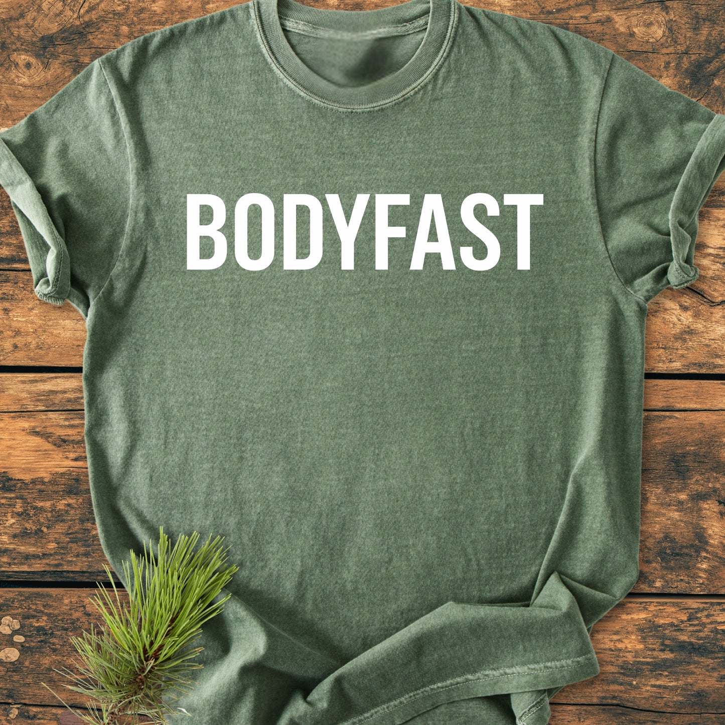 BODYFAST - Vintage Wash Tee displayed flat on wooden planks, features BODYFAST in white block letters across chest, accented with a small pine sprig, symbolizing energy and balance.