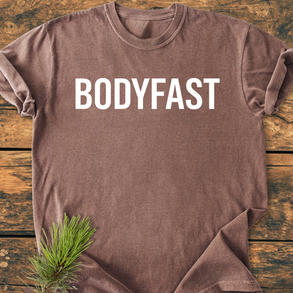 BODYFAST - Vintage Wash Tee features prominently in white letters on a brown t-shirt, laid on wood, with a pine sprig accentuating the rustic, lifestyle-inspired design.