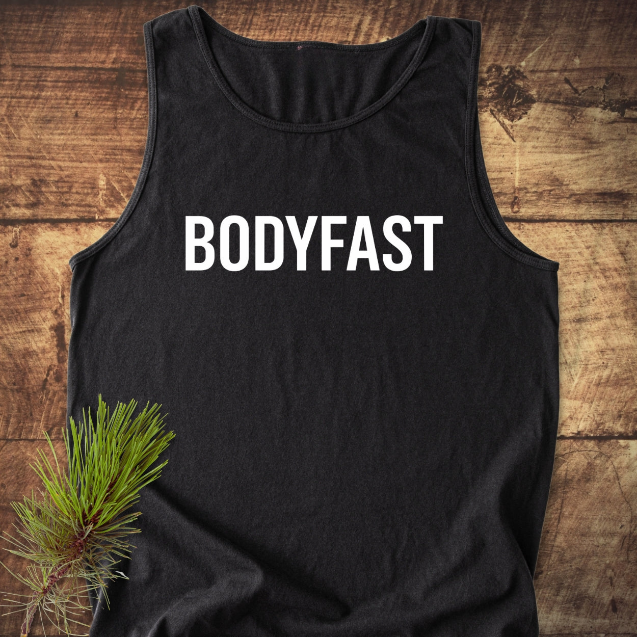 Black BODYFAST Tank Top displayed on a wooden surface, featuring bold white lettering. Accompanied by a pine branch accent, this unisex garment is crafted from sustainable, 100% ring-spun US cotton.
