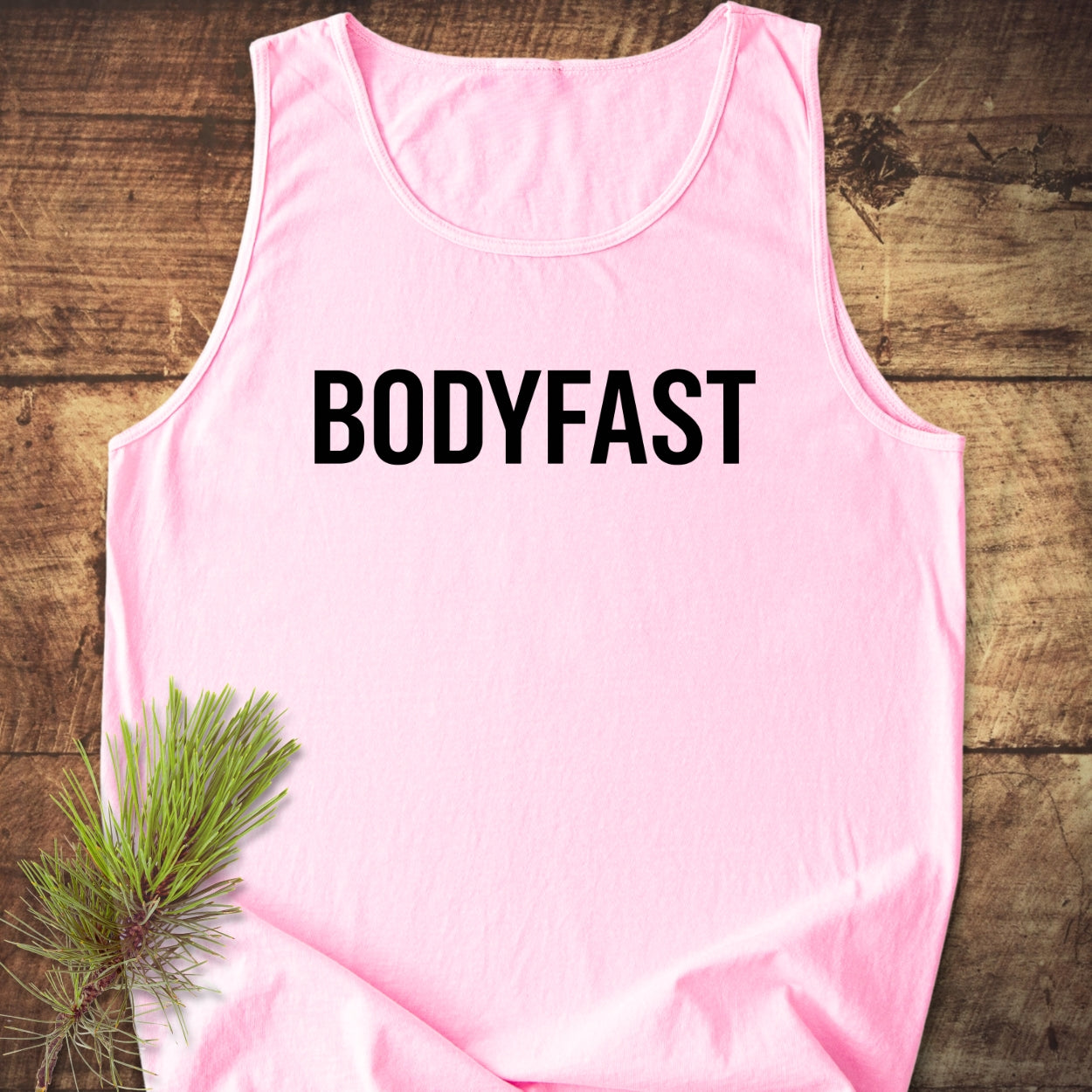 BODYFAST Tank Top in light-pink, sleeveless, laid on wood. Features bold black lettering BODYFAST across chest, with pine sprig nearby. Unisex, 100% US cotton, relaxed fit, ethically made.
