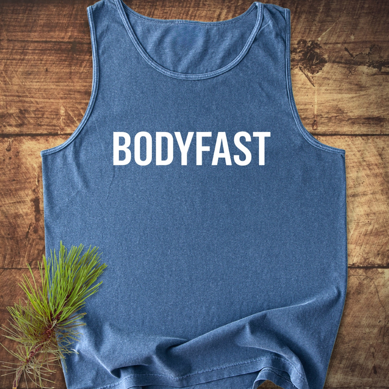 Heathered-blue BODYFAST Tank Top, sleeveless, laid flat on wood. Features bold white logo, crafted from 100% ring-spun US cotton, embodying relaxed fit and sustainability with ethical production.