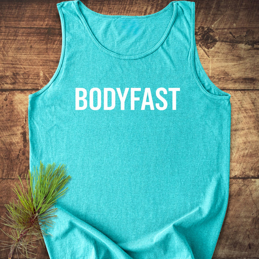 BODYFAST Tank Top, a teal sleeveless shirt with bold white lettering, lies on wood with a pine branch accent. Made from sustainable U.S. cotton, it offers a relaxed, stylish fit.