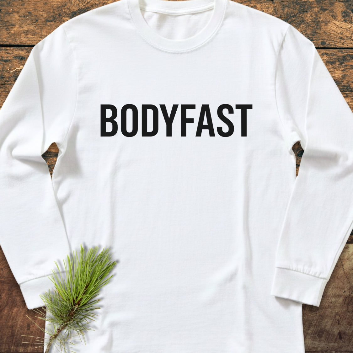 BODYFAST Long-Sleeve Tee in white, featuring bold black lettering across the chest. Laid flat with a pine sprig accent, showcasing its relaxed fit and soft, garment-dyed 100% ring-spun cotton fabric.