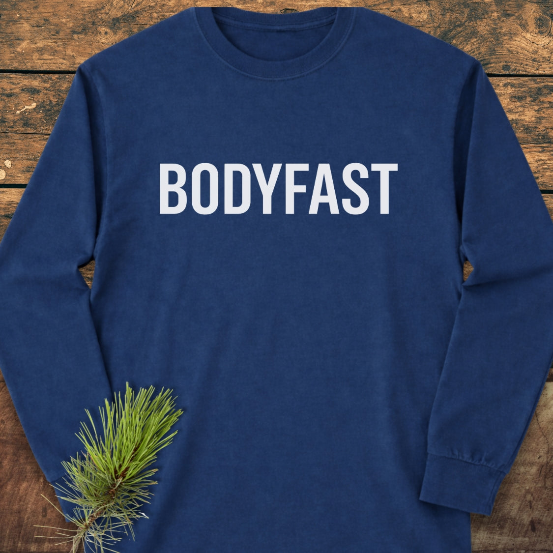 BODYFAST Long-Sleeve Tee on wooden surface, featuring large white “BODYFAST” text across the chest. Made of 100% ring-spun cotton, it offers a relaxed fit with a soft, garment-dyed fabric.