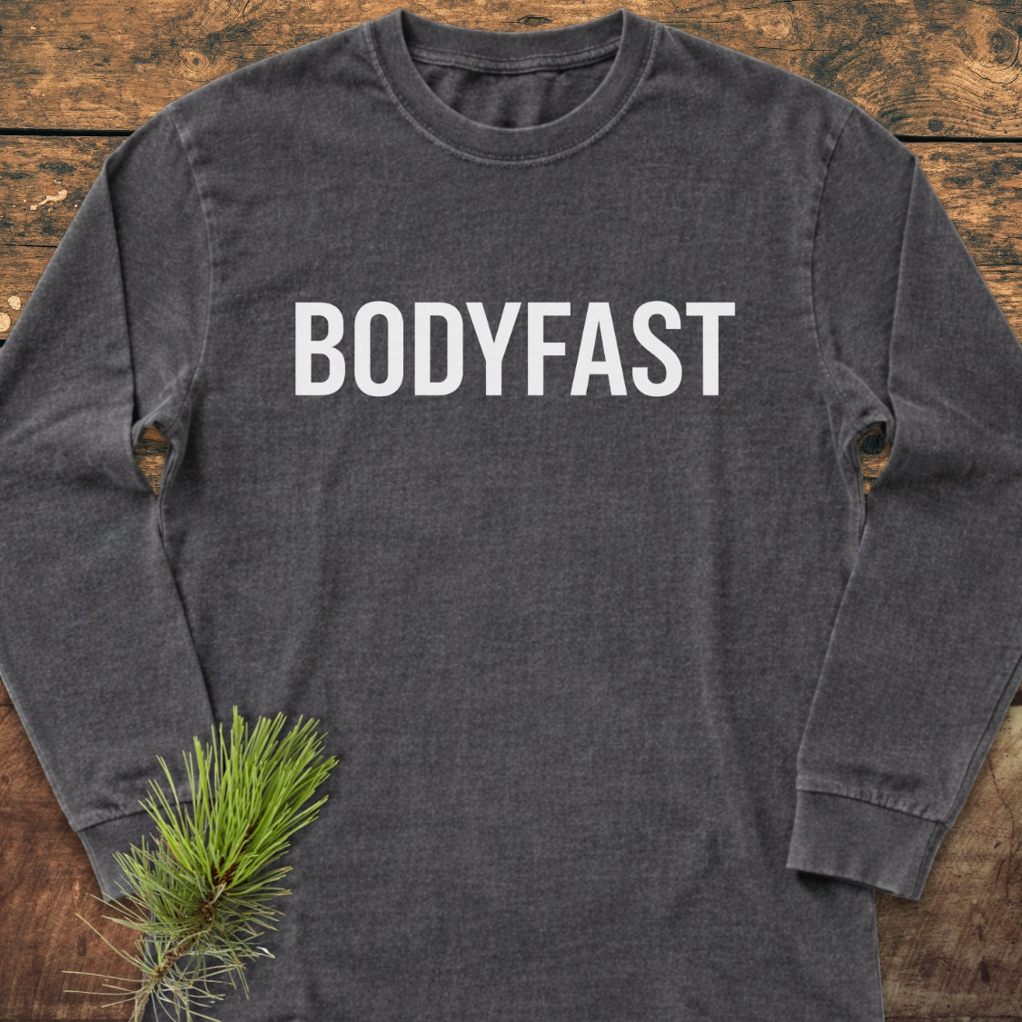 BODYFAST Long-Sleeve Tee in charcoal-gray, featuring bold white lettering across the chest, laid flat on a wooden surface with a pine branch accent. Made from 100% ring-spun cotton for softness and style.