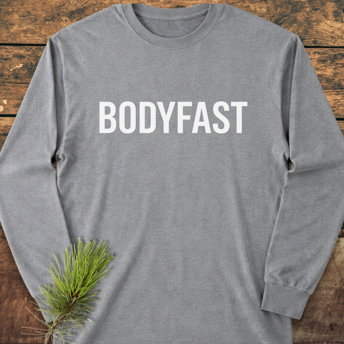 BODYFAST Long-Sleeve Tee in light-gray, featuring bold white lettering on the chest, arranged on a wooden surface with a pine sprig near the sleeve. Made of soft, 100% ring-spun cotton.
