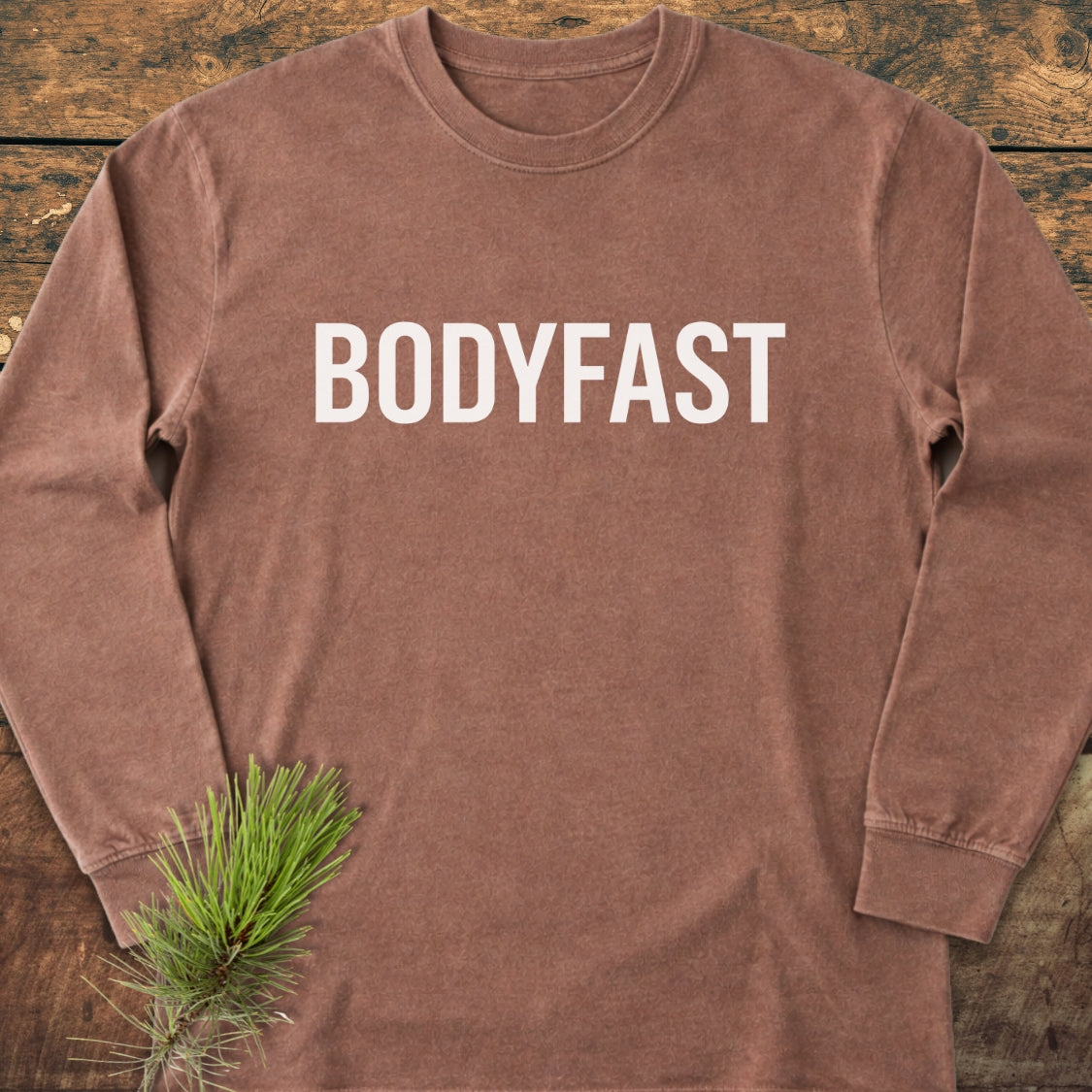 BODYFAST Long-Sleeve Tee, brown with BODYFAST in white across chest, on wooden surface with pine sprig. Made from 100% ring-spun cotton, offers garment-dyed fabric and relaxed fit.