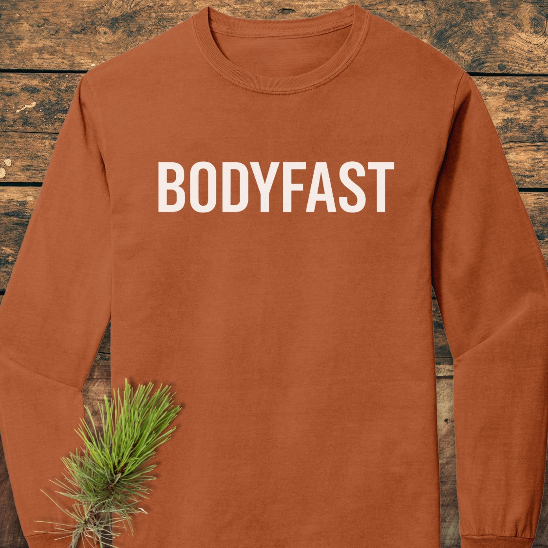 BODYFAST Long-Sleeve Tee in rust-orange displayed on wood, featuring bold white lettering. Made from soft, 100% ring-spun cotton, offering a relaxed fit and garment-dyed texture. Pine twig accessory included.