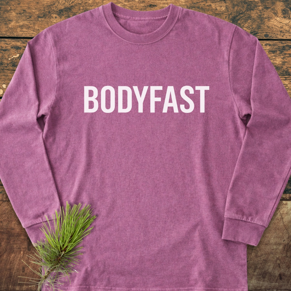 BODYFAST Long-Sleeve Tee in a heathered hue, featuring bold white lettering across the chest. Laid flat on wood, it exudes casual comfort, accented by a pine sprig.
