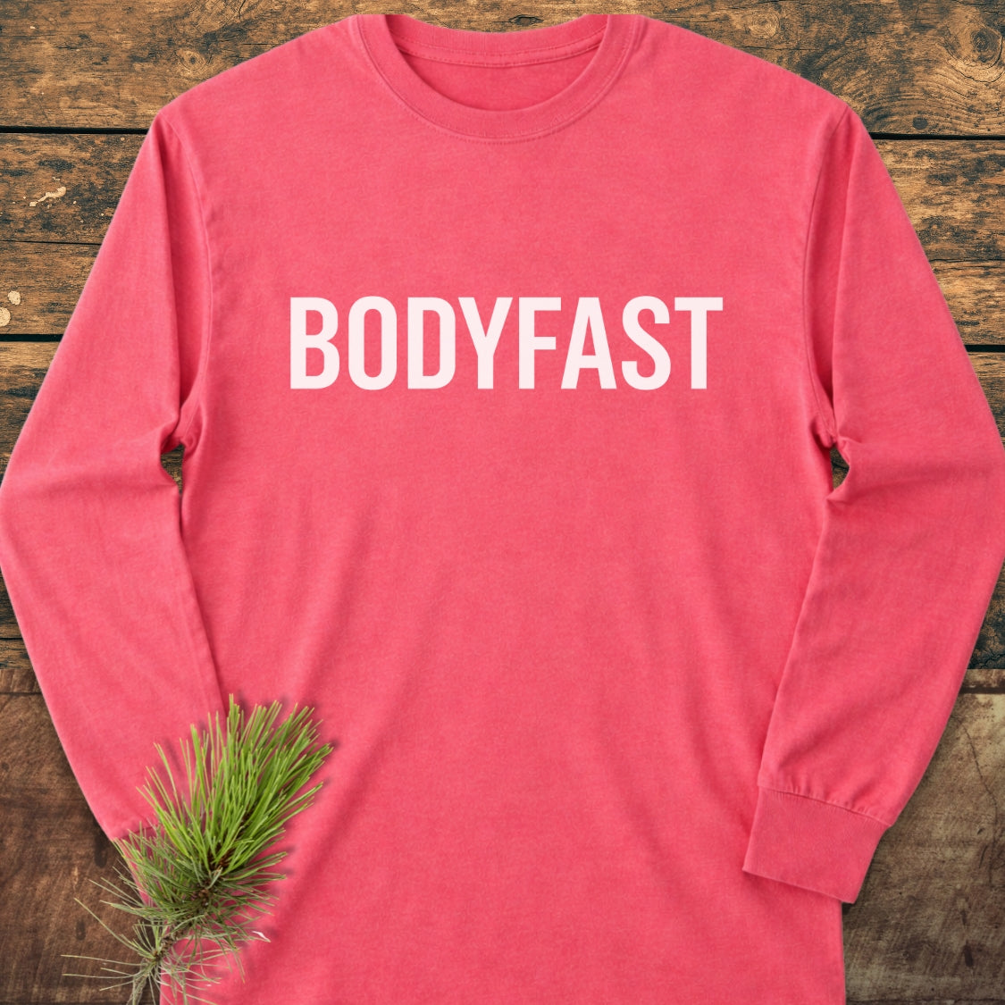 BODYFAST Long-Sleeve Tee in washed-out red, laid flat on a wooden surface, featuring bold white lettering on the chest and a pine needle sprig for a rustic touch.