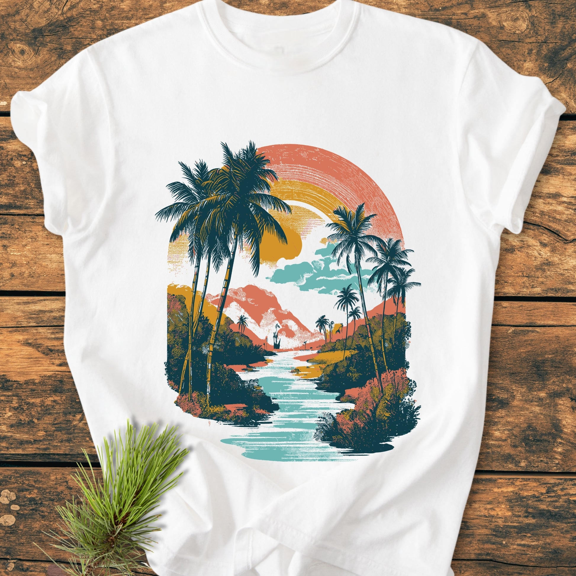 Along The River - Vintage Wash Tee features a retro landscape print with a setting sun, palm trees, and a winding river, laid flat on a wooden surface with a pine sprig accent.
