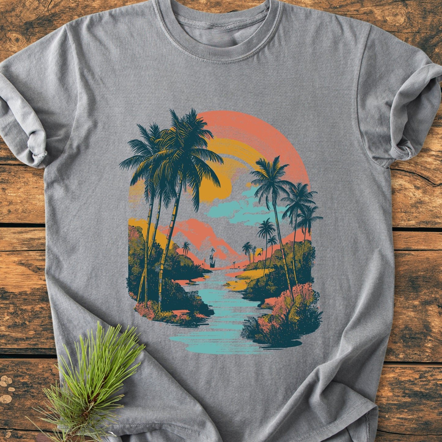 Along The River - Vintage Wash Tee features a heather-gray base with a retro tropical scene graphic, showcasing palm trees and a setting sun. Laid flat on a rustic wooden surface.