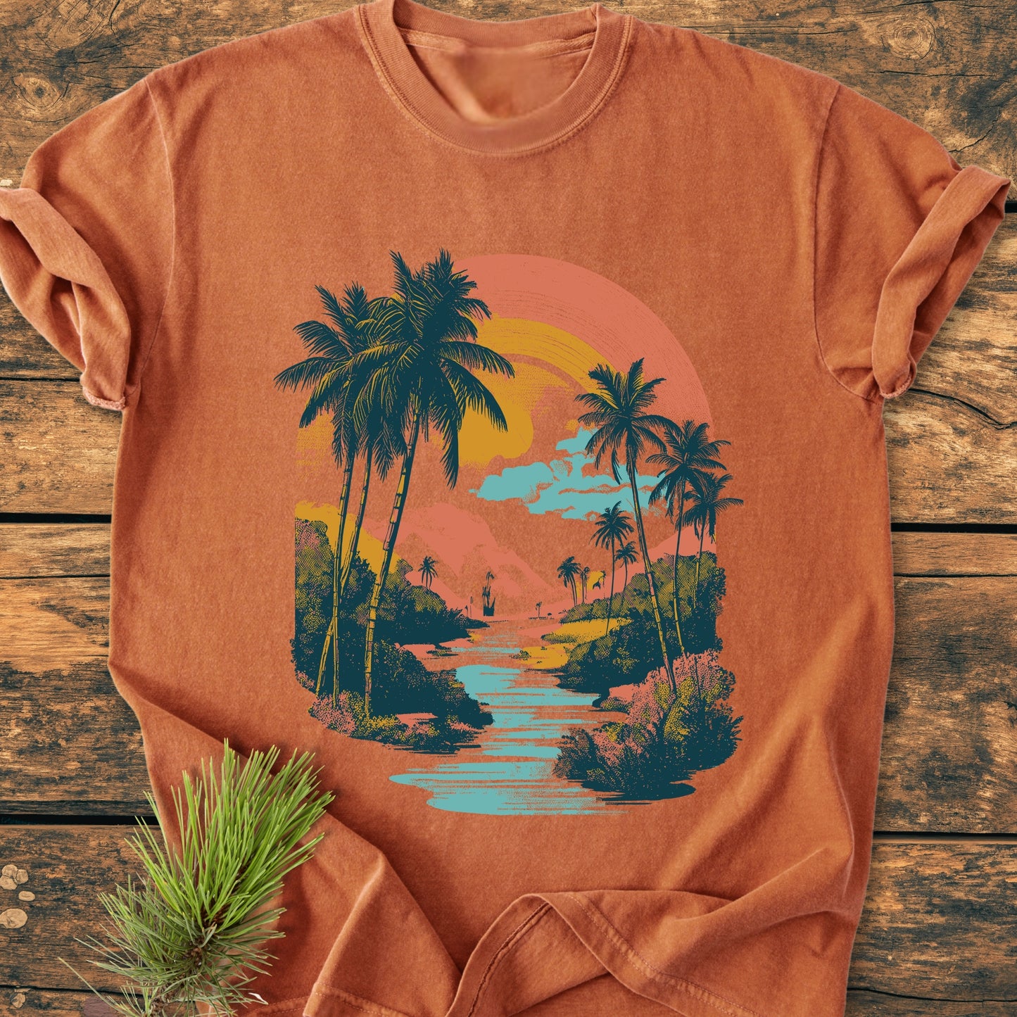 Along The River - Vintage Wash Tee features a stylized tropical sunset scene with palm trees, a winding lagoon, and distant hills, printed on soft, washed-orange fabric, laid flat on rustic wood.